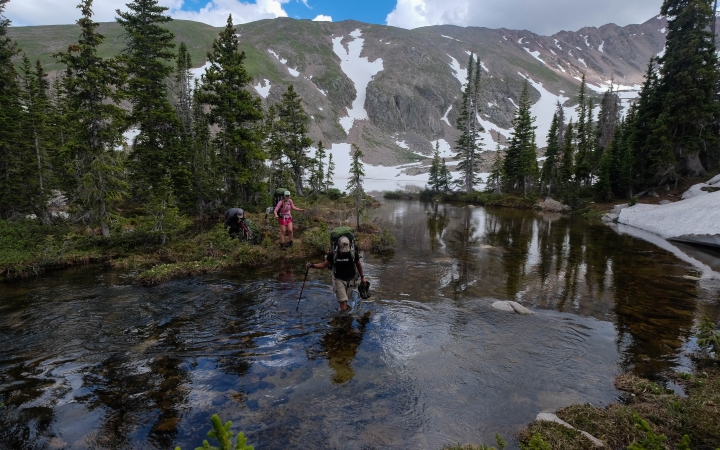 A person wades through shallow water while another person stands on the shore. Behind them are evergreen trees and mountains. 